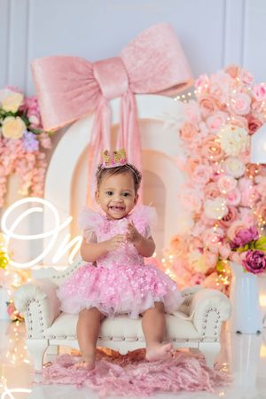 A baby girl in a pink sequin dress and crown sits on a white chair at her first birthday photoshoot with flowers.