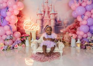 Baby girl in pink dress sits on white sofa at princess castle first birthday cake smash setup.