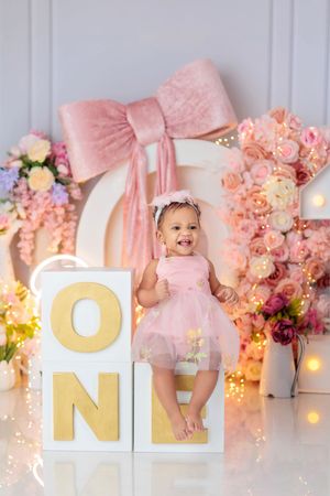 A baby girl in a pink tutu dress smiles at her first birthday photoshoot with ONE blocks and floral backdrop.