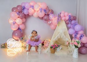 Baby in purple tutu sits before pink balloon arch with teepee, flowers, and One neon sign for birthday.