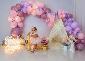 Baby in pink tutu sits by balloon arch, floral teepee, and One neon sign for first birthday photoshoot.