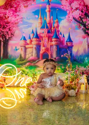 Baby girl in gold dress sits before a Disney castle backdrop with pink trees and a neon 'One' sign.
