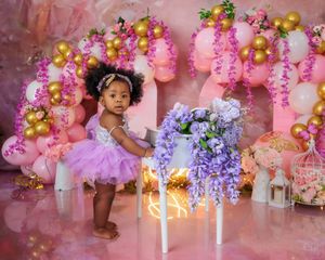 Baby girl in lavender dress stands by floral table with pink balloons and gold accents on pink backdrop.