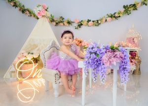 A baby girl in a purple tutu sits on a white chair at her first birthday photoshoot with purple floral decorations.