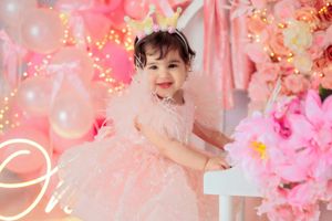 Smiling baby girl in pink feathered dress sits at white piano with pink floral balloon backdrop.