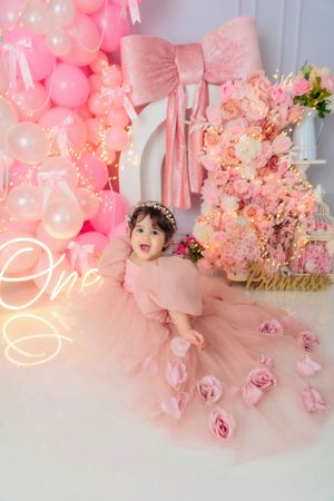 Baby girl in pink ball gown sits on floor with One neon sign and pink balloon backdrop.