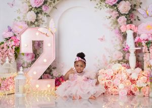 Baby girl in pink dress sits before floral arch with illuminated number 2 and roses backdrop.
