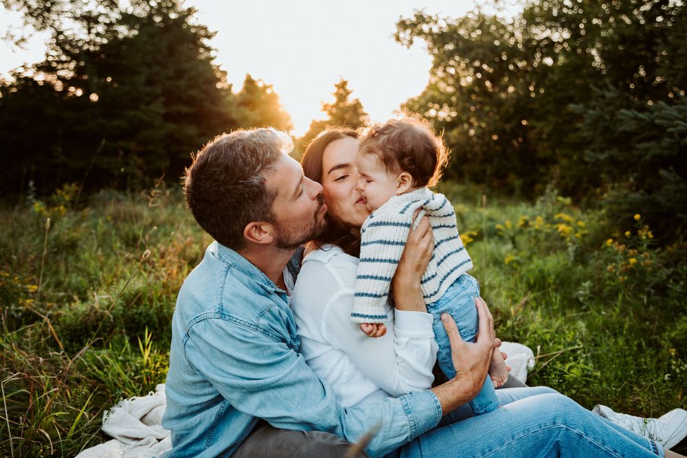 Family sharing kisses while sitting on blanket in grassy field during sunset.