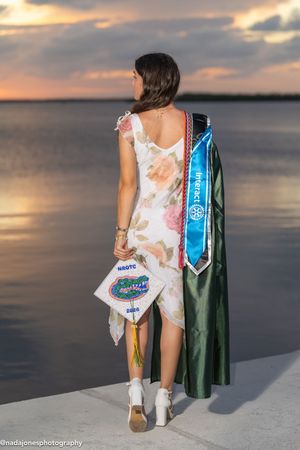 Woman in floral dress holding graduation cap and sashes poses by waterfront at sunset.