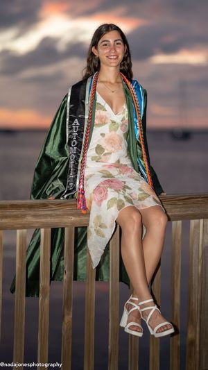 Smiling graduate in floral dress and green graduation gown sits on wooden railing at sunset with colorful honor cords.