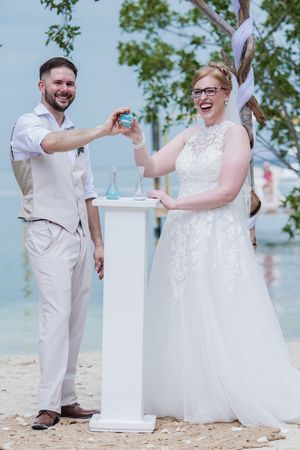 Happy couple performing a sand ceremony on a tropical beach wedding, bride in white lace gown and groom in tan suit.