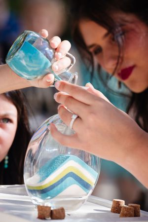 Woman carefully pouring colorful layered sand into a glass vase, creating a decorative sand art display.