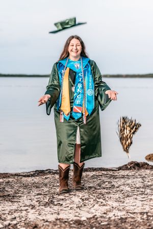 A smiling graduate in green robe and blue sash tosses cap near waterfront wearing cowboy boots.