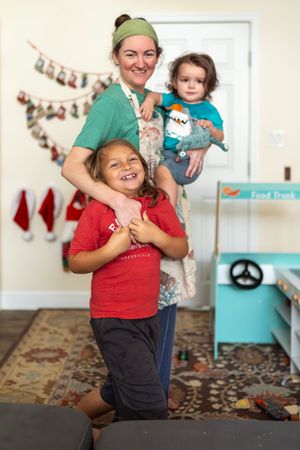 A smiling woman in green holds a toddler while an older child hugs her waist indoors.