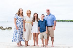 Family of five smiling together on a sandy beach, dressed in blue and white outfits with calm ocean waters behind them.