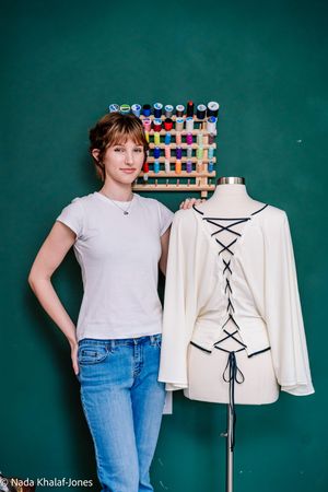 Young woman in white t-shirt and jeans stands beside a dress form displaying a cream lace-back blouse in a sewing studio.