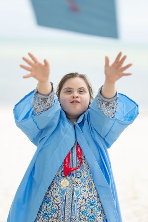 Young girl with Down syndrome in blue raincoat joyfully raises hands outdoors near beach.