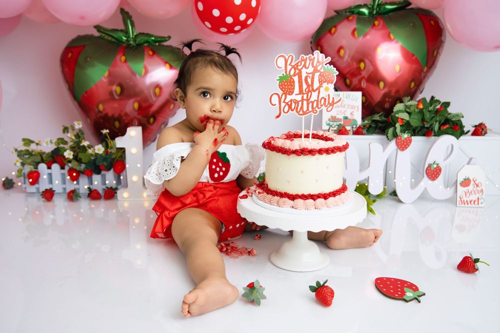 Festive strawberry-themed cake smash photography setup with red balloons and white cake on pink background.