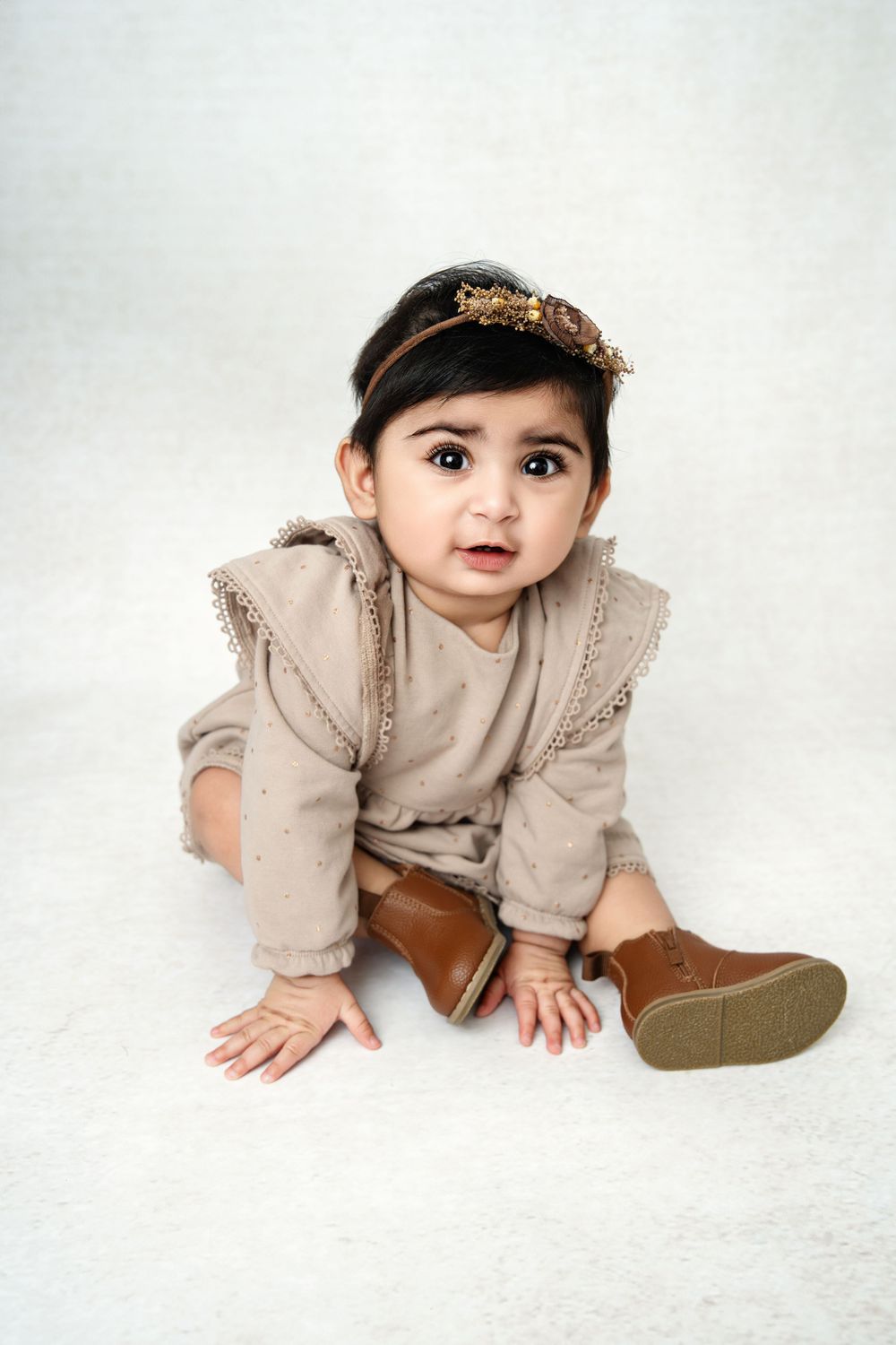 Baby in beige outfit crawls on white backdrop during professional photo session.