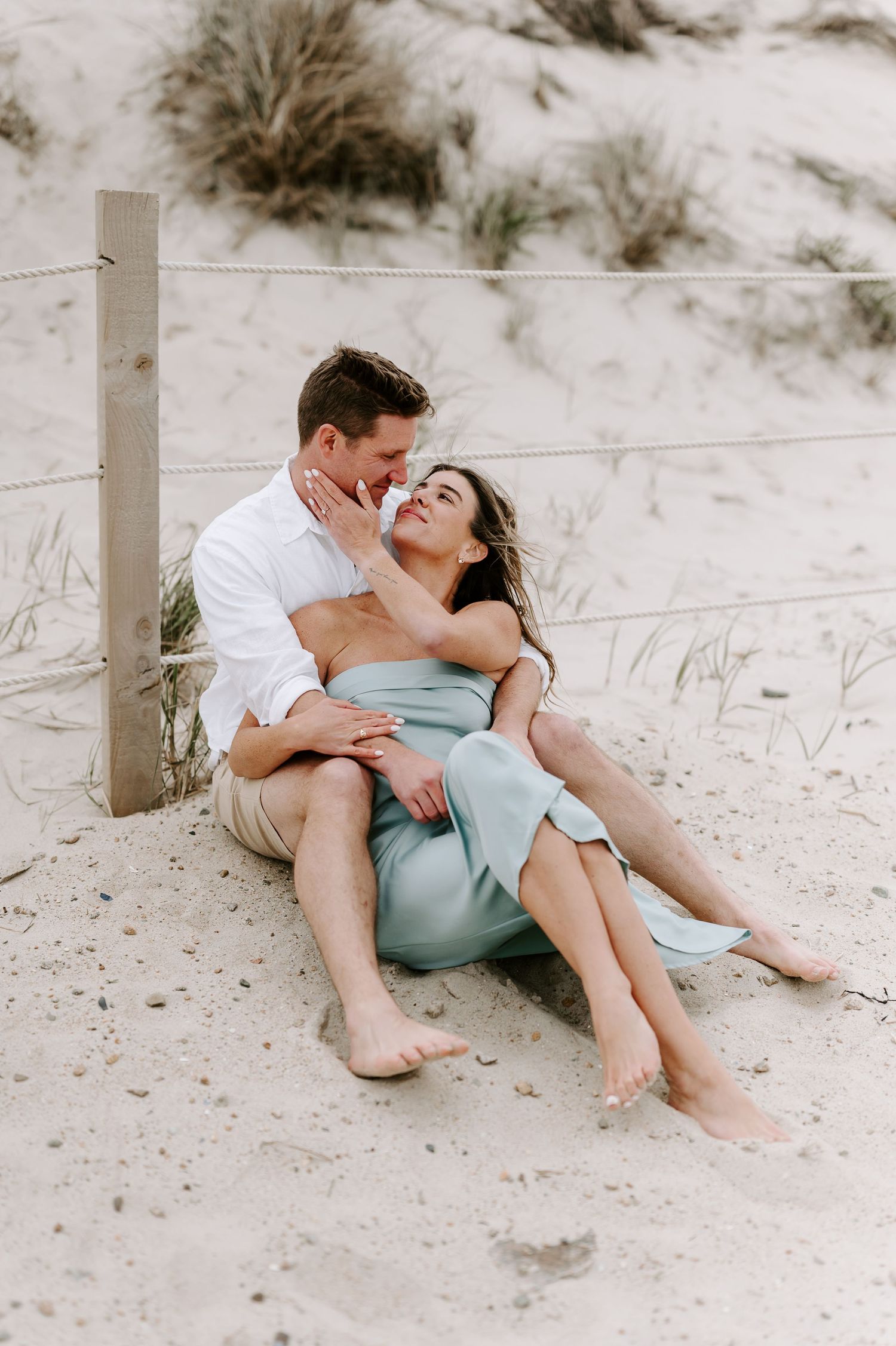 A couple shares an intimate moment while sitting in sandy dunes near a wooden post.