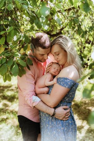 Family portrait under green foliage as parents embrace their newborn in natural lighting.