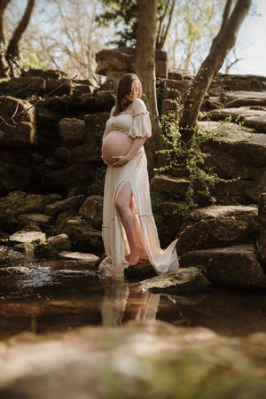 Romantic maternity photo by a creek with white dress and natural stone backdrop.