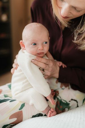 A baby in white clothing rests on floral bedding while being gently held taken by photographer pausing time photography