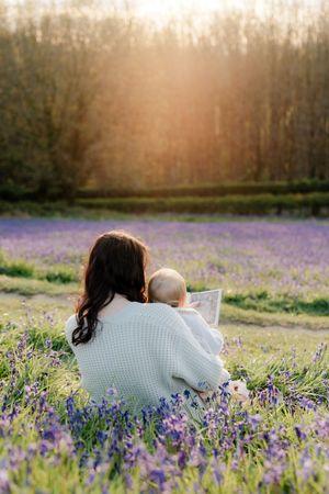 Mother sitting in purple lavender field holding baby reading a book at golden sunset hour.