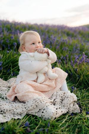 Baby girl in white sweater sits on lace blanket in lavender field holding stuffed bunny at sunset.