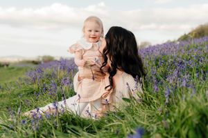 A dark-haired mother sits in a bluebell field holding her smiling baby girl on a bright spring day.