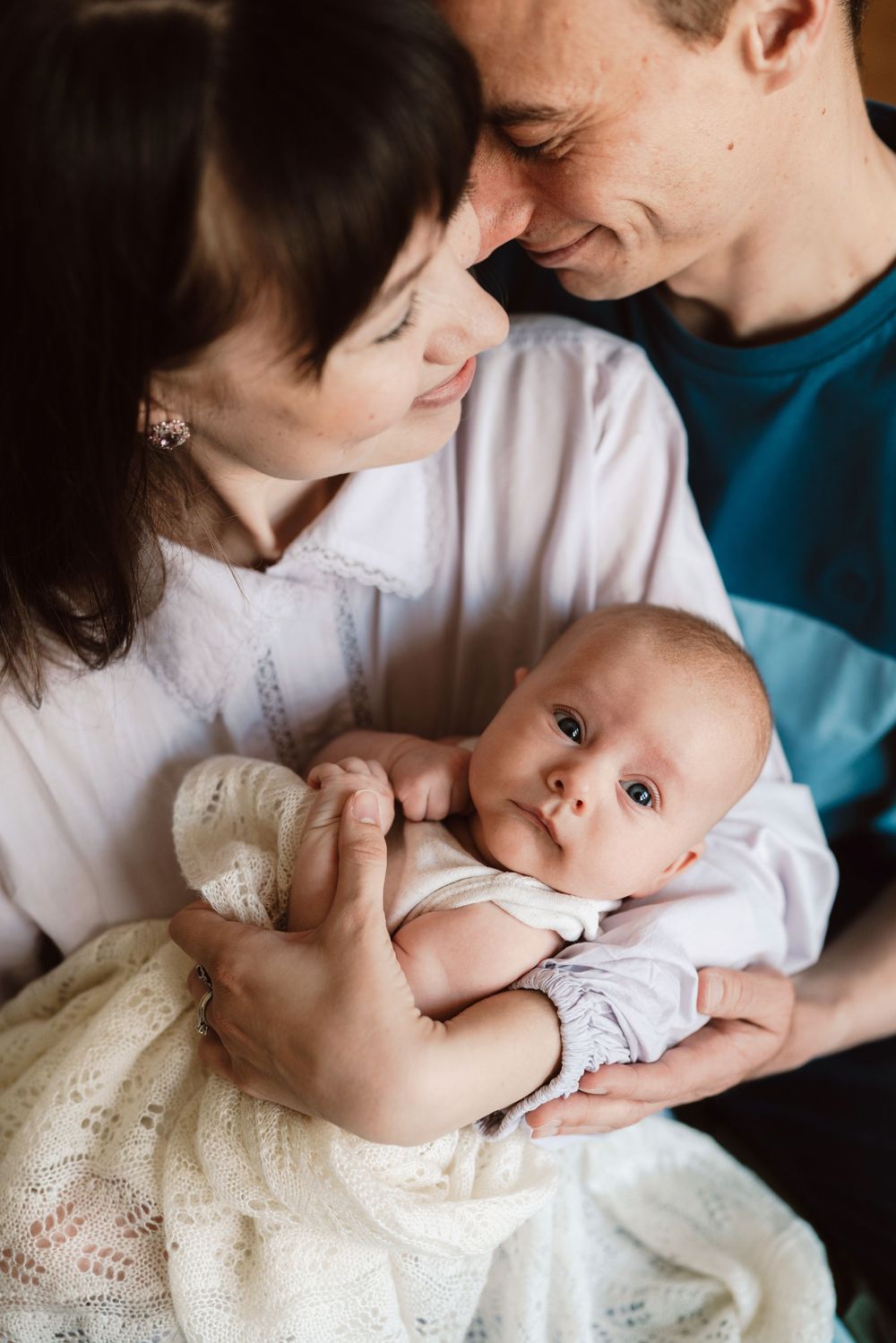 Happy parents smiling at each other while lovingly holding their newborn baby dressed in white.