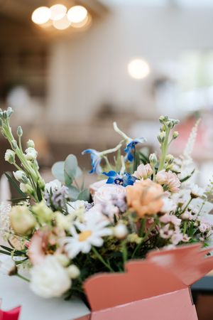 A vibrant mixed flower bouquet with blue, pink, and white blooms in a pink box, set against a softly blurred interior.