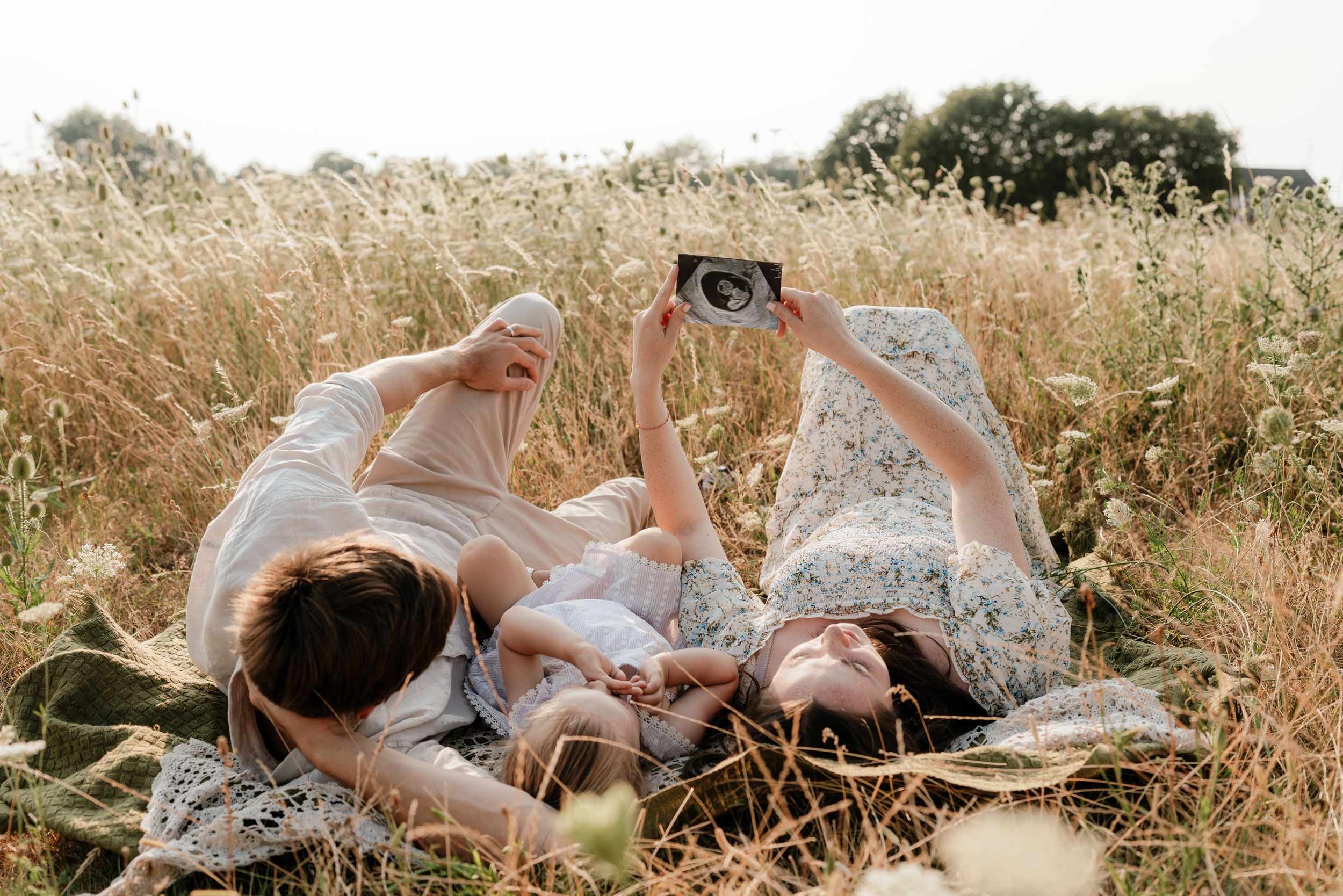 Three people lying in a golden field holding an ultrasound photo, celebrating a pregnancy announcement outdoors.