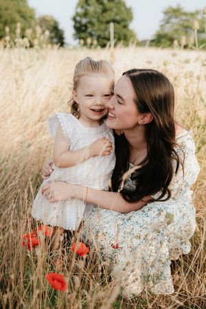 A mother in a floral dress kisses her smiling toddler daughter in a golden field with red poppies.