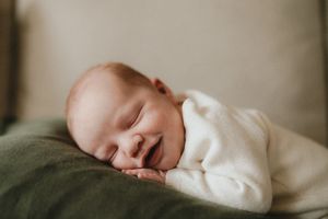 Smiling sleeping newborn baby in white onesie resting on green blanket in soft natural light.