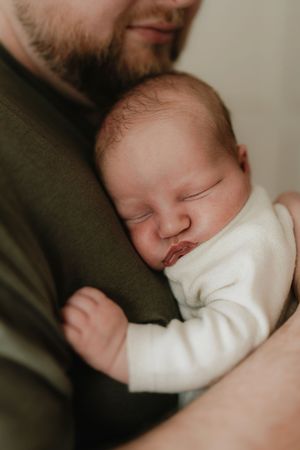 A newborn baby sleeping peacefully in a father's arms, dressed in a white onesie.