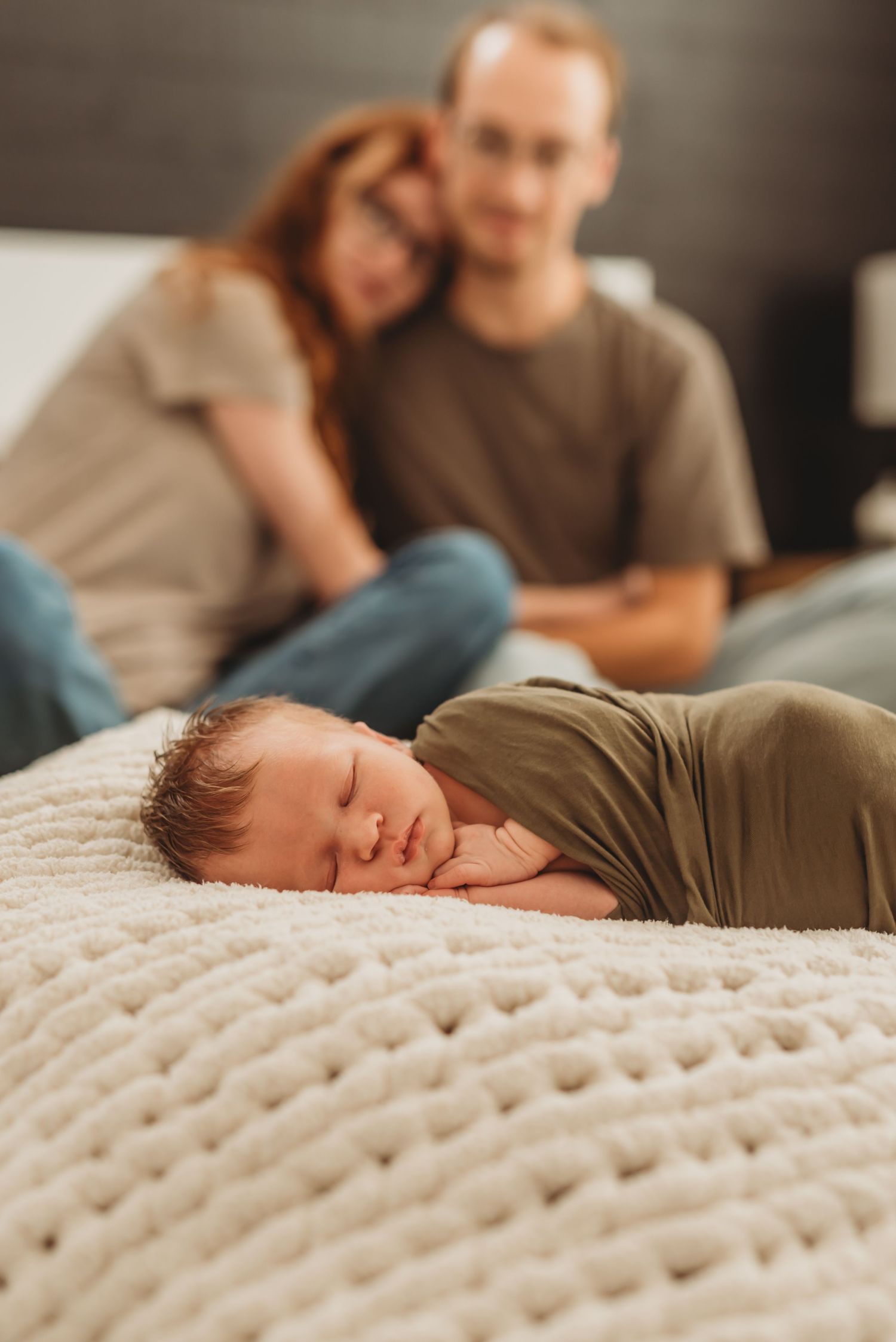 Baby sleeping peacefully on chunky knit blanket while parents relax in background.