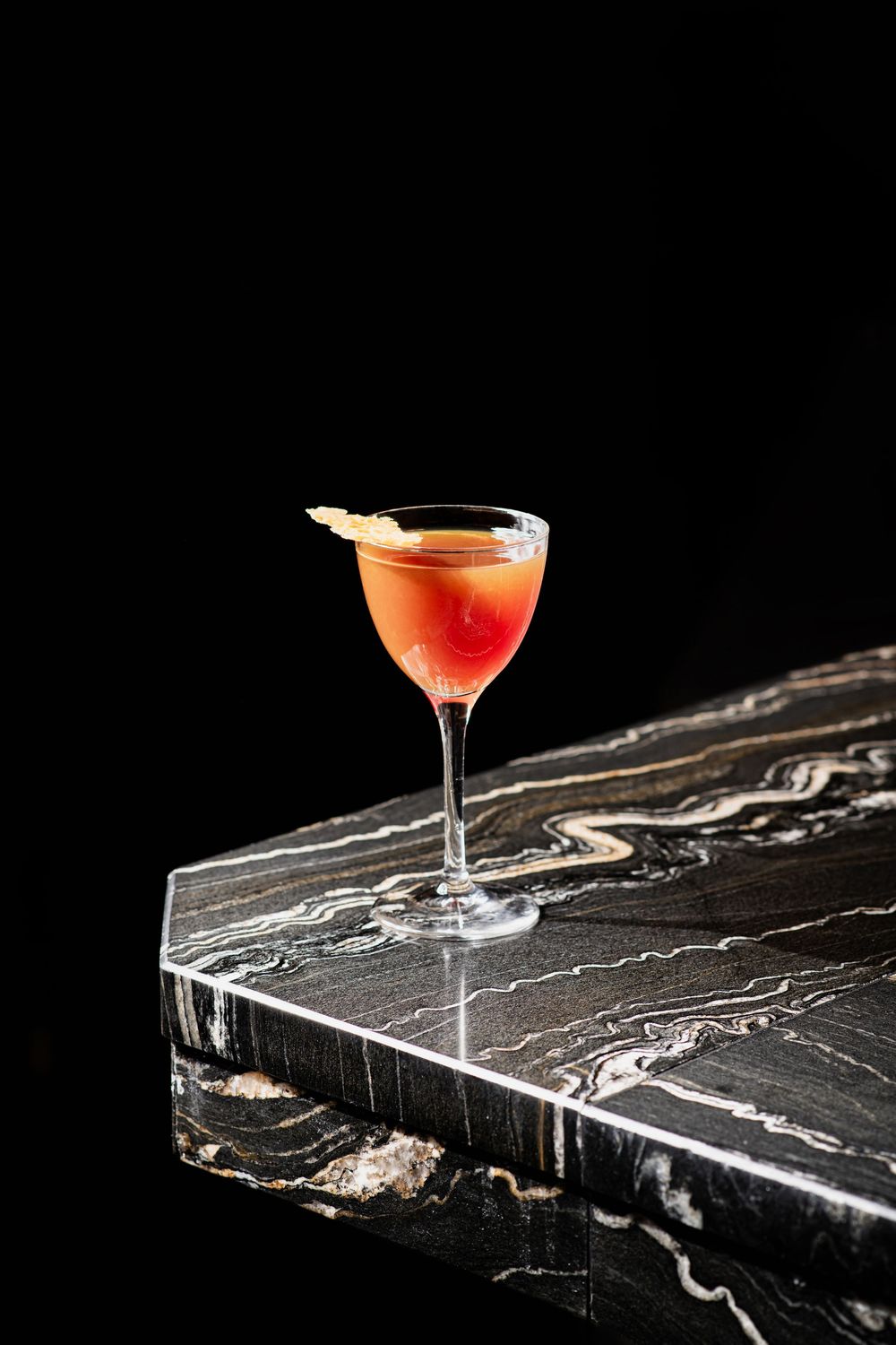 Red cocktail in a coupe glass on black marble surface with dramatic lighting.