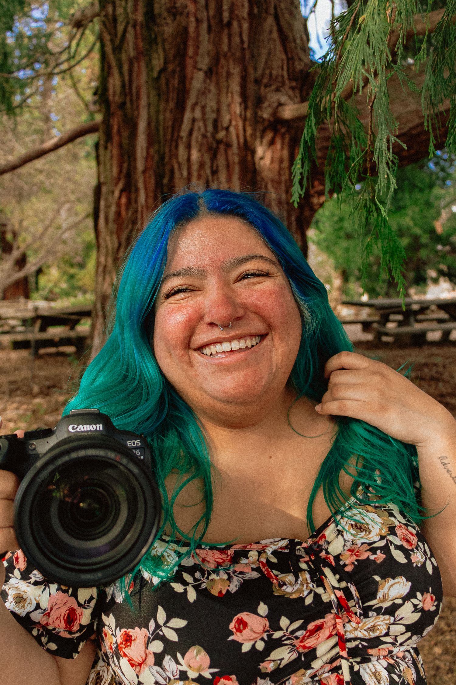 A Mexican woman photographer in a floral dress and teal hair holds a camera.