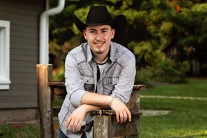 A person in a black cowboy hat leans on a wooden fence post in a rural outdoor setting.