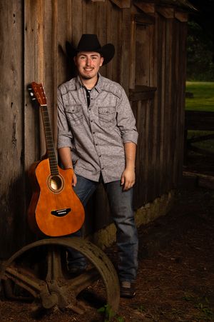 A country music performer in a cowboy hat poses with an acoustic guitar against a rustic wooden barn backdrop.