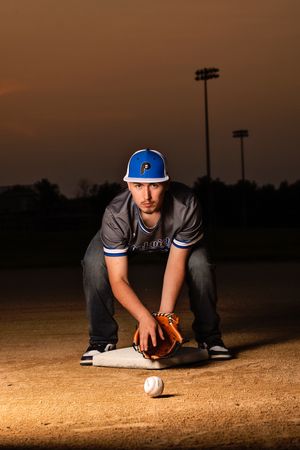 Baseball player crouches on field at dusk with ball and glove as stadium lights glow in background.