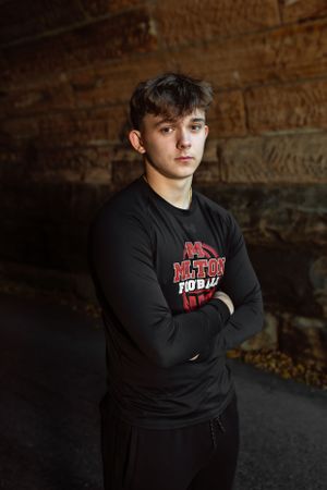 Person wearing black sweatshirt with red text logo stands with crossed arms against rustic wall in dramatic lighting. Janesville, WI
Brodhea