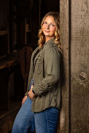 A person in an olive green button-down shirt and blue jeans poses against a rustic wooden wall in warm lighting.