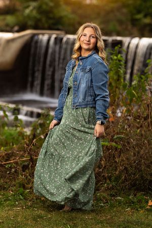 A green polka dot dress paired with a denim jacket is worn in a natural outdoor setting with a waterfall backdrop.