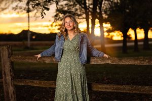 A person in a floral dress poses in a golden sunset at a rustic farm setting with wooden fences and trees.
