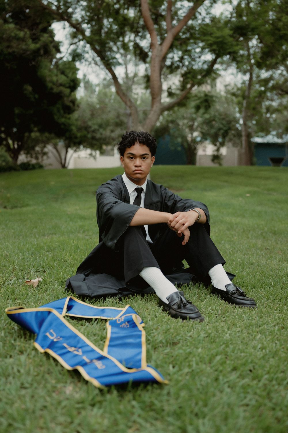 A graduate in formal attire sits on grass with a blue graduation banner lying beside them in a park setting.