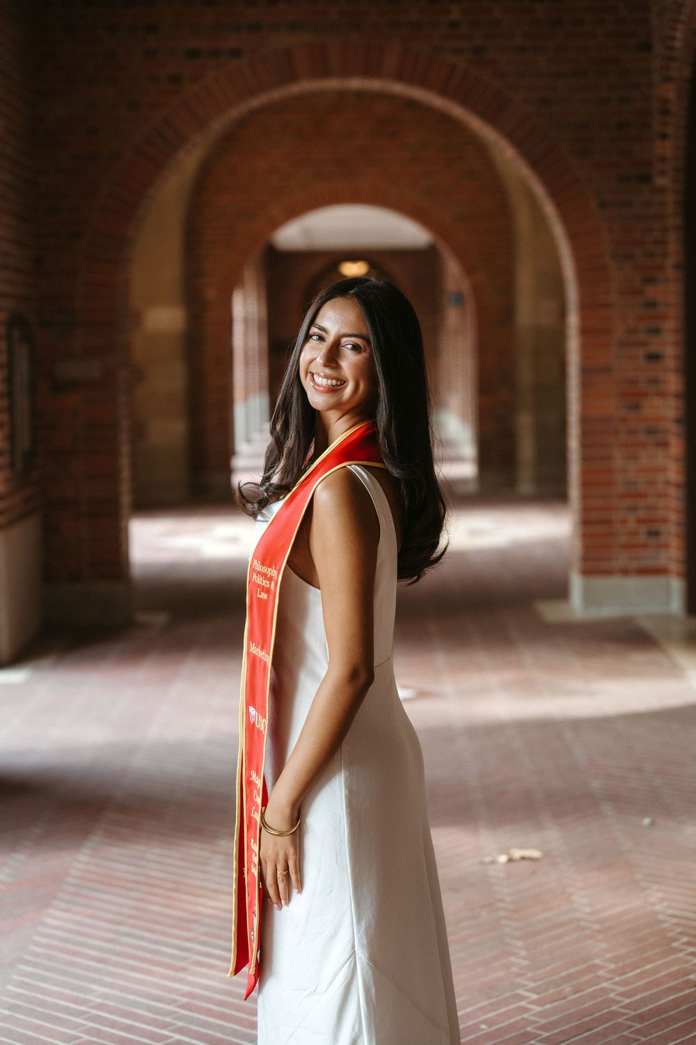 A woman in a white dress and red stole poses in a brick archway corridor with dramatic lighting.