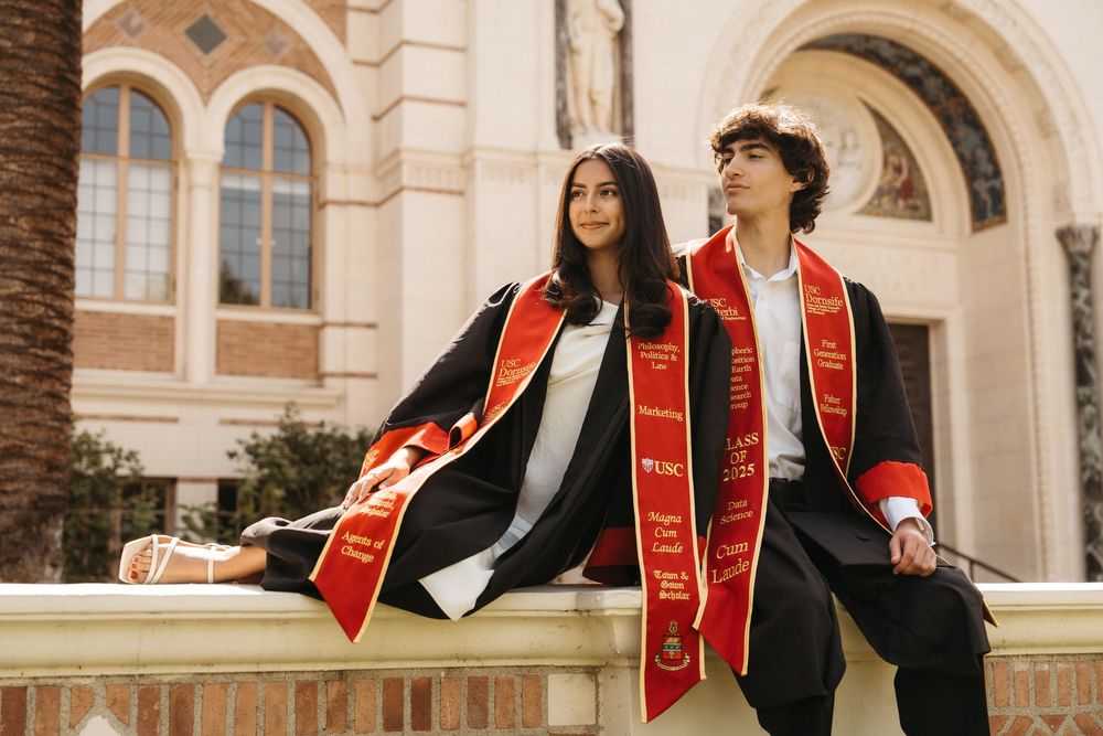 College graduates in black caps and gowns with red stoles celebrate on campus steps.
