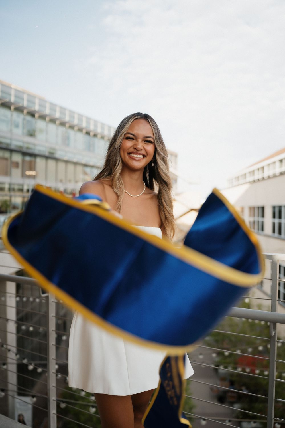 A graduate in white waves a blue and gold graduation stole against an urban backdrop.